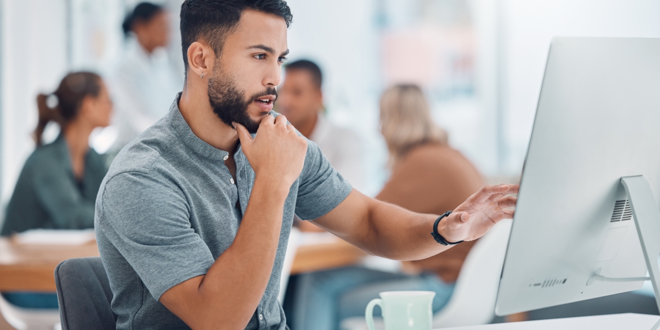 Dark haired man with beard wearing a grey short sleeved shirt looks at a computer monitor while sat in the office