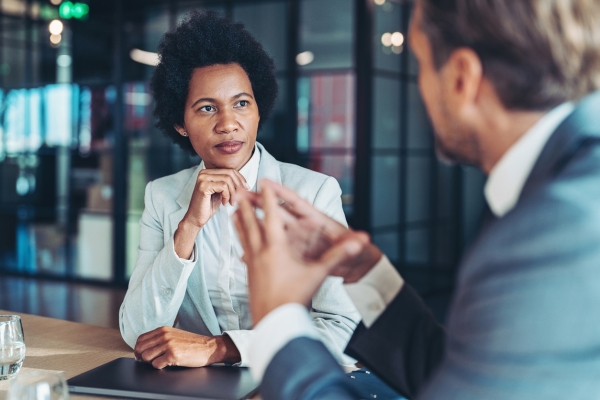 A smartly dressed Black businesswoman is sat at a table with her hand on her chin. She is listening to a white man dressed in a suit sat to her left