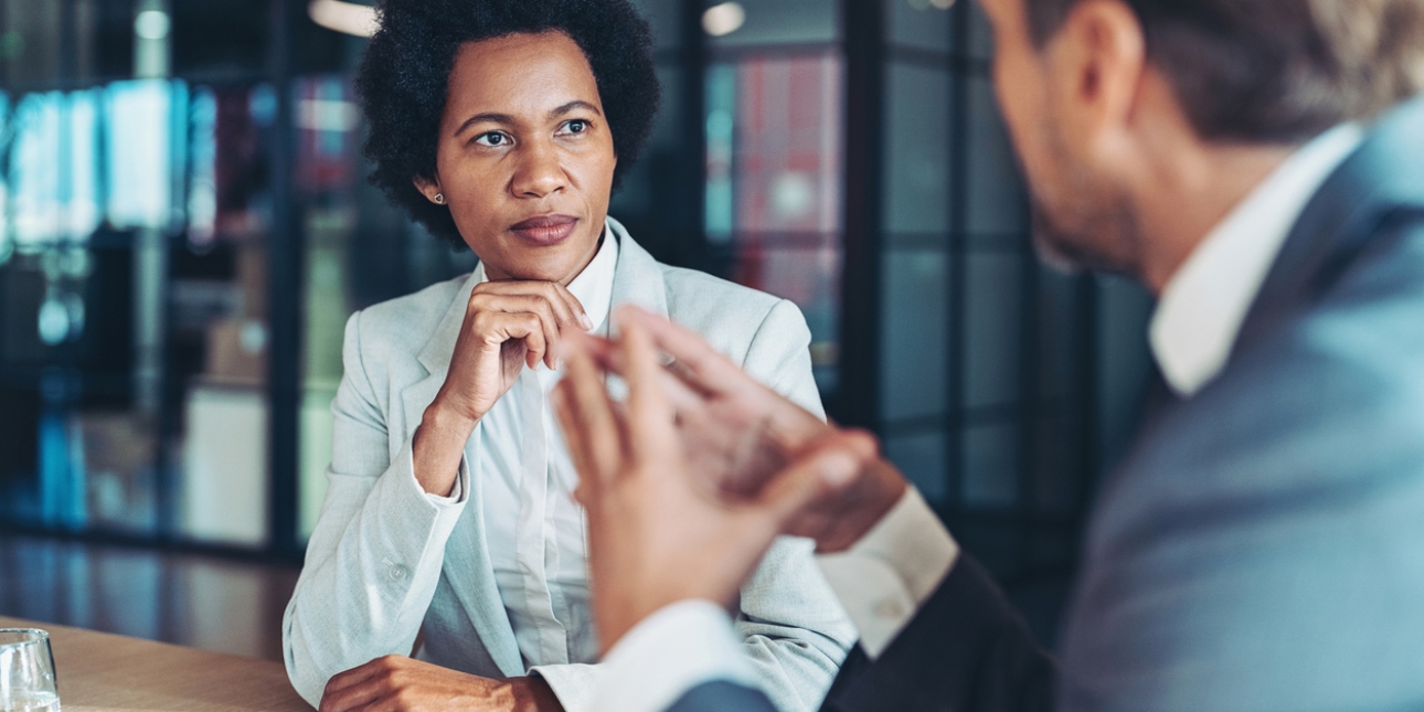 A smartly dressed Black businesswoman is sat at a table with her hand on her chin. She is listening to a white man dressed in a suit sat to her left