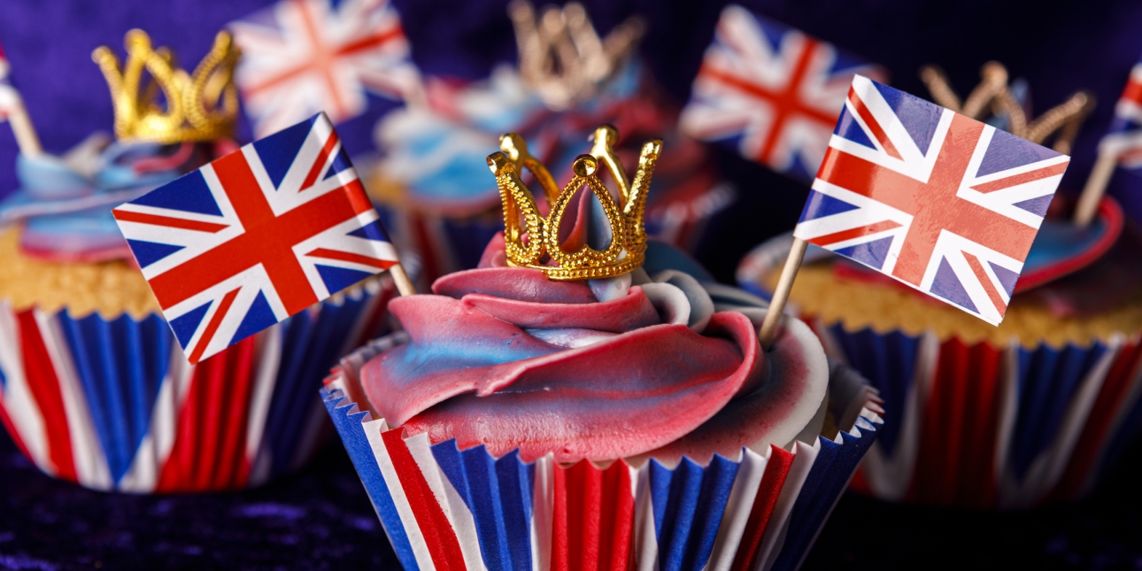 A selection of red, white and blue cupcakes with miniature gold crowns on top. There are assorted miniature two union (jack) flags stuck in each cake.