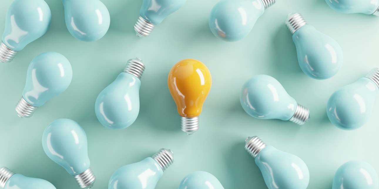 A bird's eye view of a yellow tinted light bulb laid flat on a teal background, surrounded by 15 turquoise-tinted bulbs