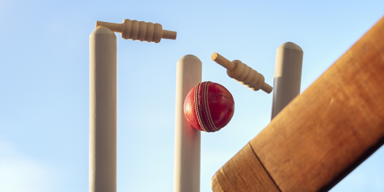 The lower part of a cricket bat is visible in front of a cricket ball which has knocked two bails from the cricket stumps