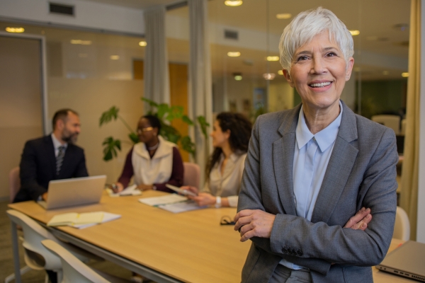 A white middle aged woman with grey hair, wearing a grey suit, stands with her arms cross in a boardroom. A white man, black woman and white woman are sat in the background.