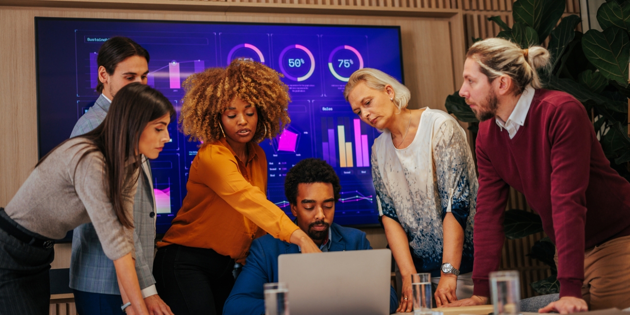 A multi-ethnic group of four colleagues are stood looking at the laptop of another colleague who is sat at a table. In the background is a screen displaying graphs and charts.