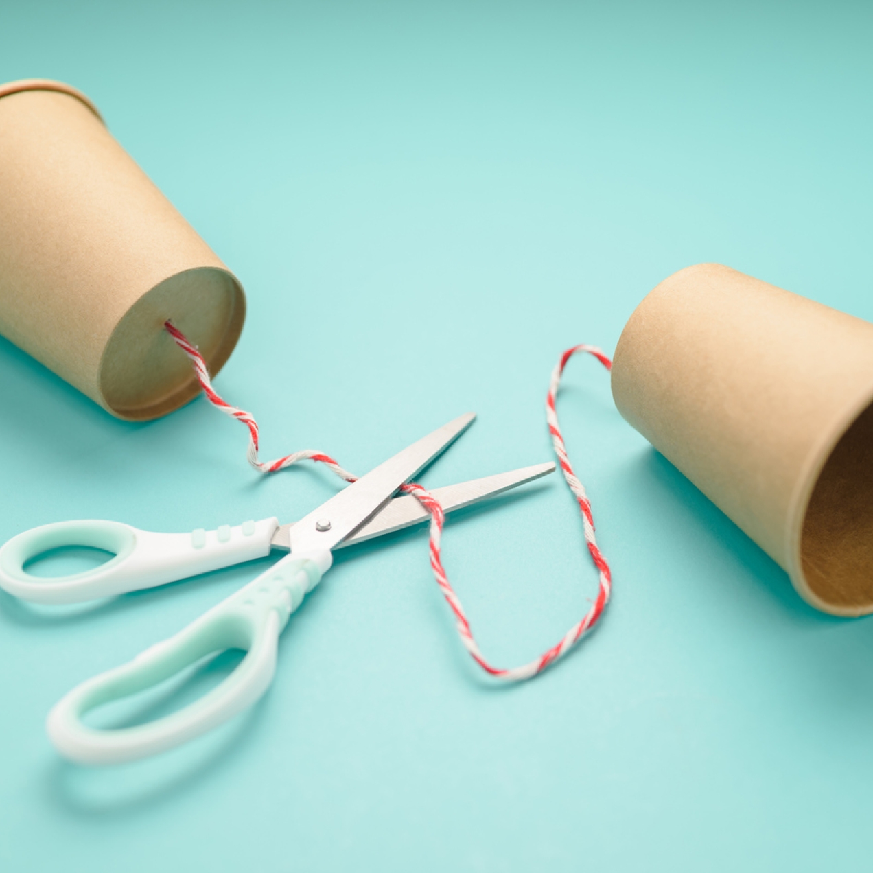 Two brown paper cups connected by string through a hole in the bottom of the cups. A pair of scissors is open and ready to cut the string.