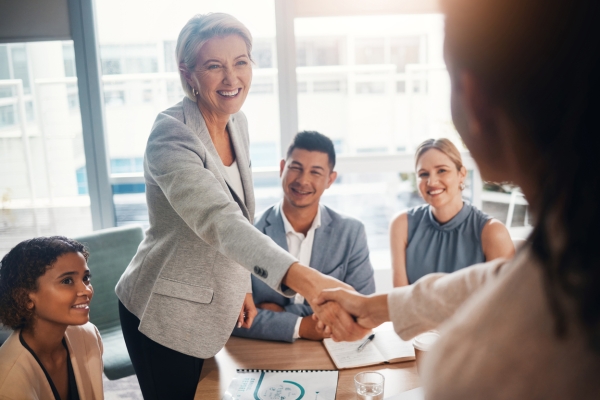 A smartly dressed woman with a welcoming smile, stands at a board table shaking her hands with another woman who is out of focus. Around the table are sat two other women and one man who are all smiling and wearing business attire.