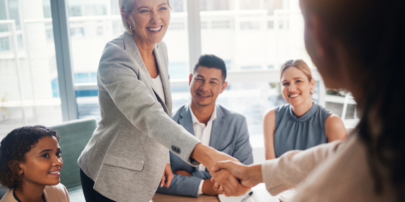 A smartly dressed woman with a welcoming smile, stands at a board table shaking her hands with another woman who is out of focus. Around the table are sat two other women and one man who are all smiling and wearing business attire.