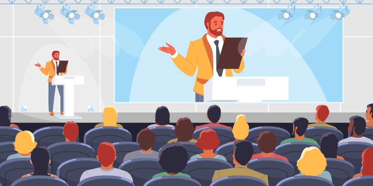 Illustration of a man with a beard and wearing a yellow blazer stood on stage. His image is projected on to a large screen next to him. In the foreground are the backs of audience members seated in four rows.
