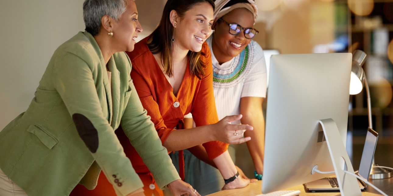 Three women of different ages and ethnicities lean over a desk looking at a computer monitor