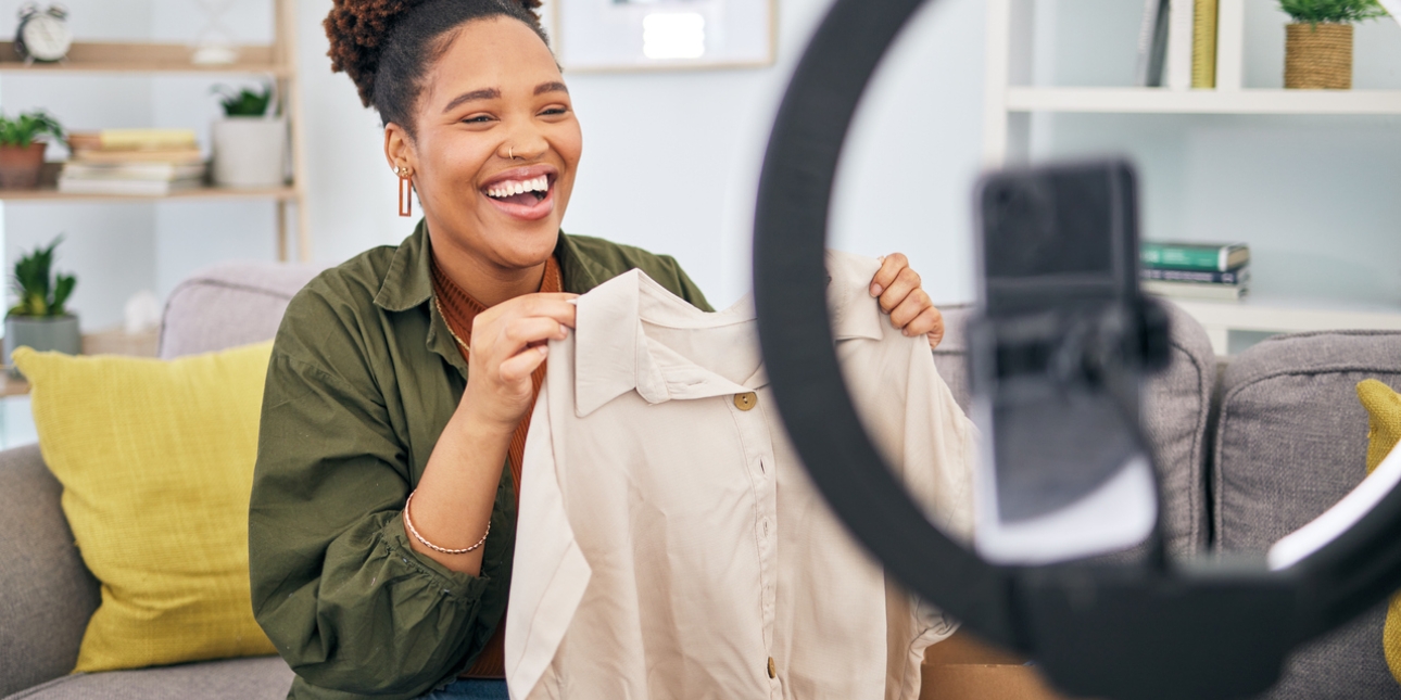 A black woman, who is sat on a sofa, laughs while holding a jacket in front of a mobile phone and ring light. In the background are a cushion and plants, books and a clock on a wooden shelving unit.