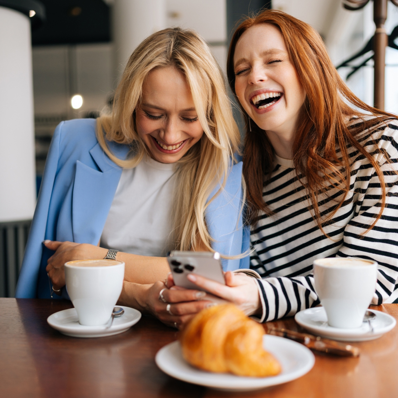 Two women, both white - one with blonde hair, the other with red hair, laughing while looking at a mobile phone screen. They are sat in a café.