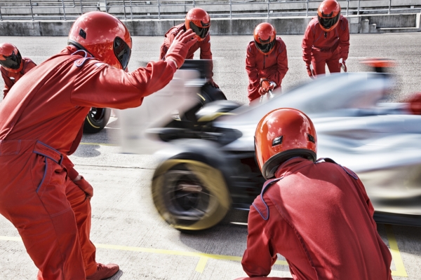 A team of six racing engineers, dressed in red overalls and red crash helmets with visors pulled down, wave off a racing car from the pit stop