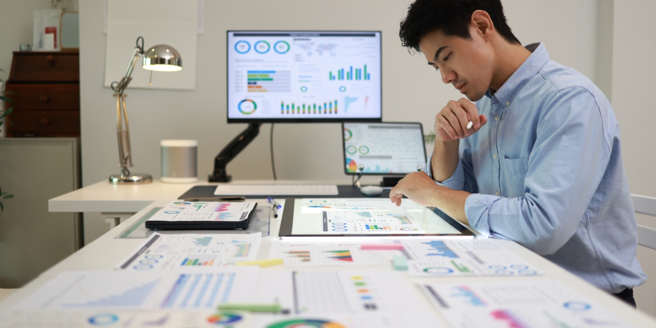 Side profile of an Asian man with short dark hair and blue shirt, sat at a desk which is covered in print outs of graphs and tables. Screens in the background show similar information, alongside a lamp and other office furniture