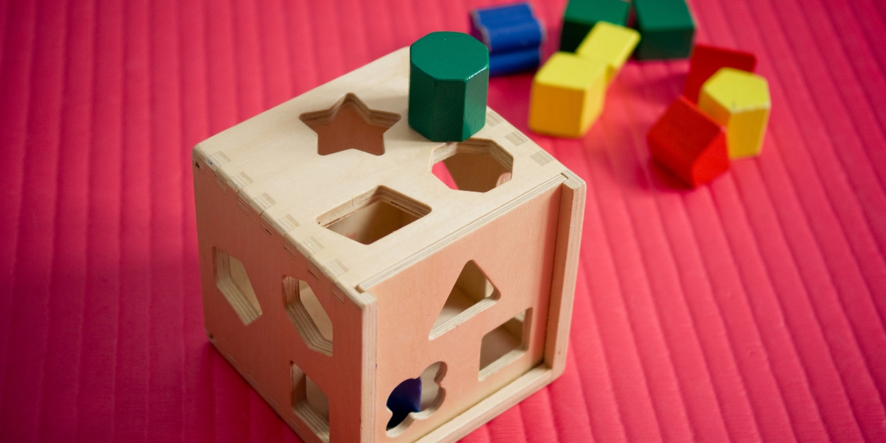 A wooden shape sorter toy with various shapes cut out of it. A an octagonal wooden block is placed on the top in between the octagonal and star-shaped holes. Other assorted blocks are scattered behind the toy.