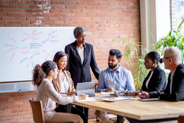 A meeting of six colleagues in business dress around a table. A Black woman manager with short hair is stood at the head of the table. On her left is a young Asian man with a beard and an older white man with grey hair. On her right is an Asian woman