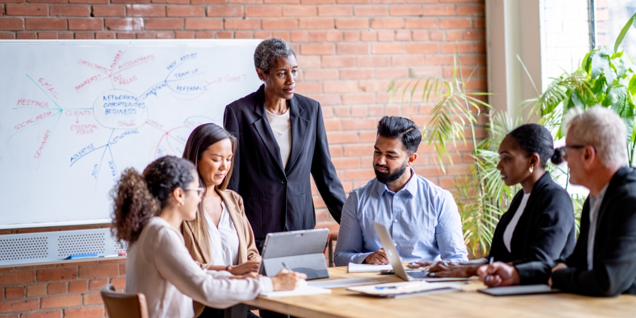 A meeting of six colleagues in business dress around a table. A Black woman manager with short hair is stood at the head of the table. On her left is a young Asian man with a beard and an older white man with grey hair. On her right is an Asian woman
