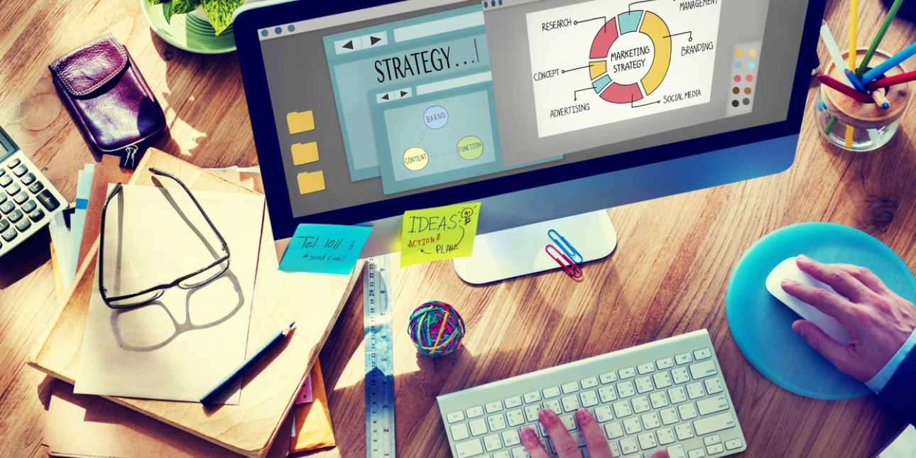 Overview of a desk with assorted stationery, spectacles and calculator. A computer screen shows an image of a marketing strategy pie chart with segments showing content management, branding, social media, advertising, concept and research. Two white