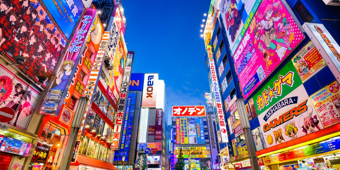 Crowds pass below brightly lit colourful signs attached to buildings of several stories in Akihabara at dusk under a dark blue sky.