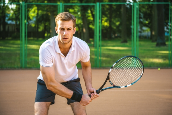 A white tennis player wearing a white shirt and black shorts grips a tennis racquet in both hands ready for the serve. In the background are a bitumen tennis court, tennis ball, fence and greenery