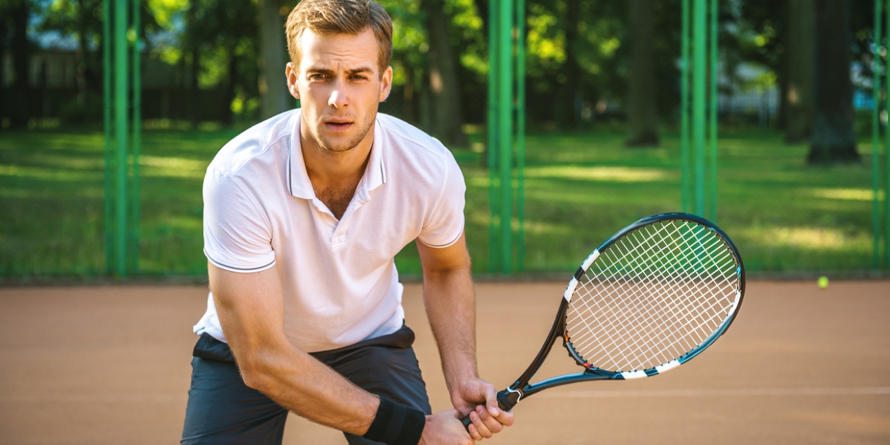 A white tennis player wearing a white shirt and black shorts grips a tennis racquet in both hands ready for the serve. In the background are a bitumen tennis court, tennis ball, fence and greenery