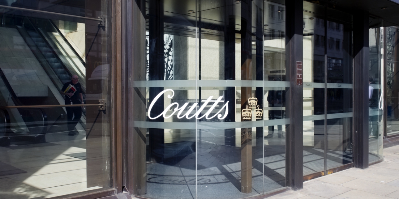 The glass entrance of Coutts bank branch on the Strand in London. The Coutts logo, compromised of the name in italic serif font and three crowns, is on the glass.