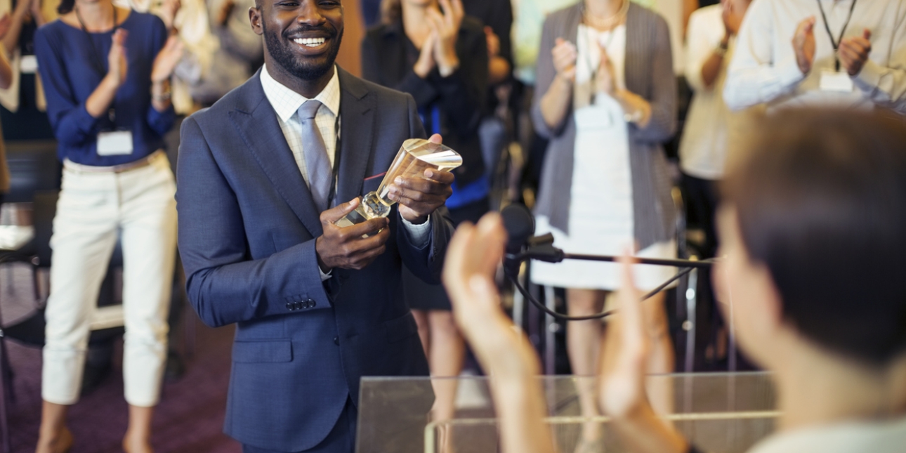 A smiling man, who is black and wearing a navy suit and light blue tie, holds a trophy in his hand. He receives a standing ovation from the audience in the background. In the foreground is the back of the out of focus head of the person giving the aw