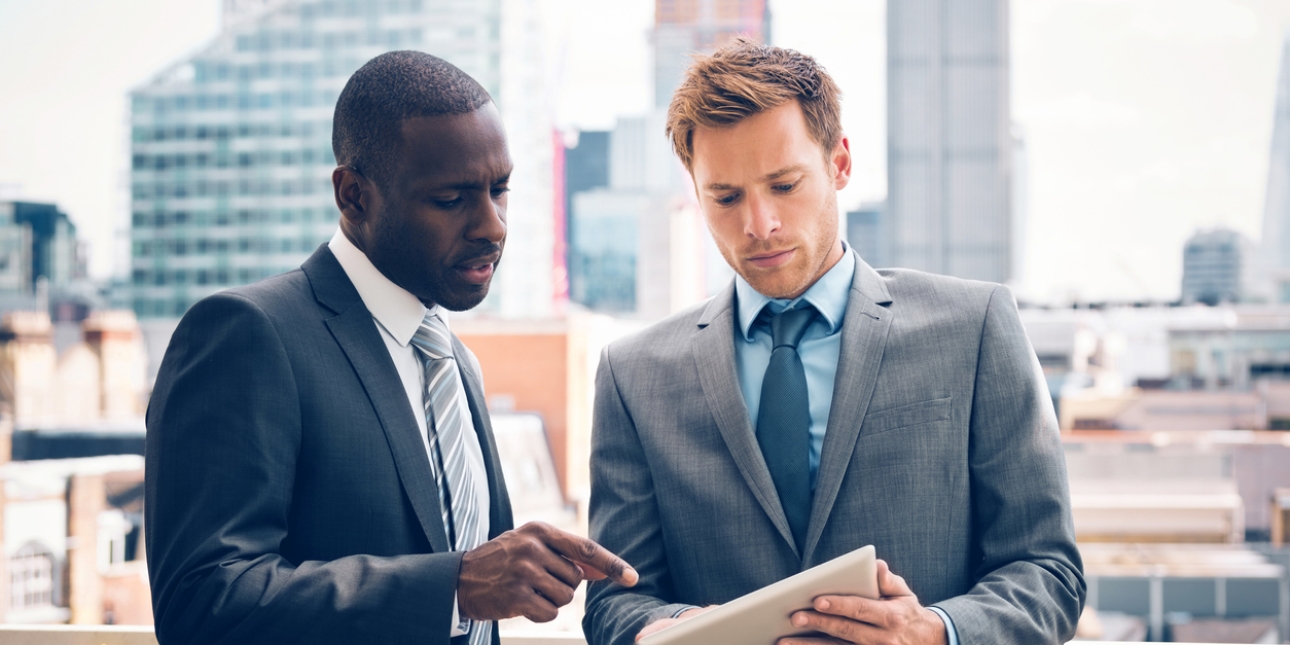 A Black man in a suit points to a tablet held by a white man in a suit. They are stood on on a balcony with the City of London skyline behind them.