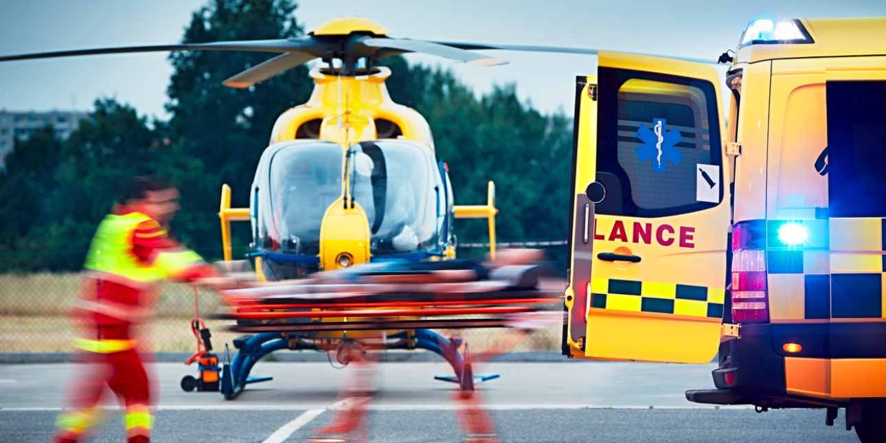 Side profile of a paramedic pushing a stretcher into the back of an ambulance. Behind them is stood an emergency helicopter.