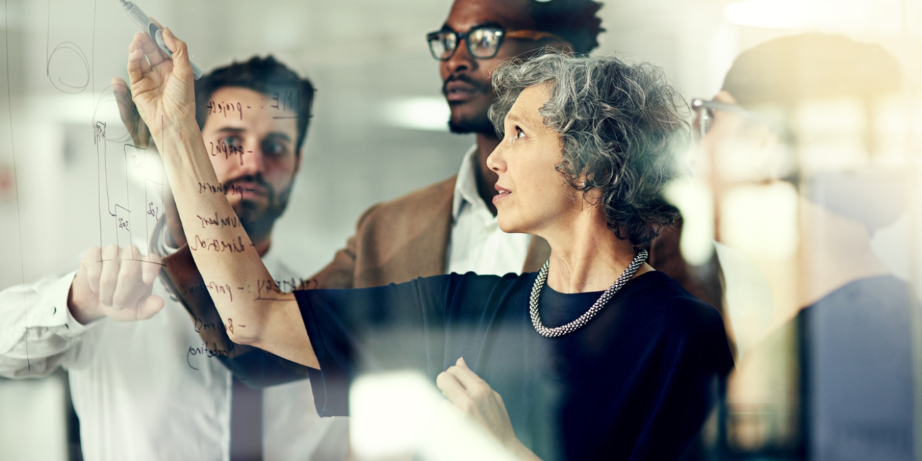 A business woman writes on glass with a marker while three male colleagues look on