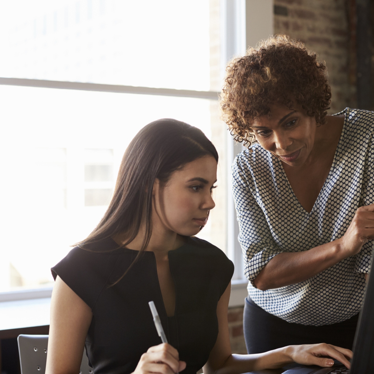 A younger woman with a black top and dark hair is sat at a computer screen. Leaning over her and also looking at the screen is an older woman wearing a spotted blouse.
