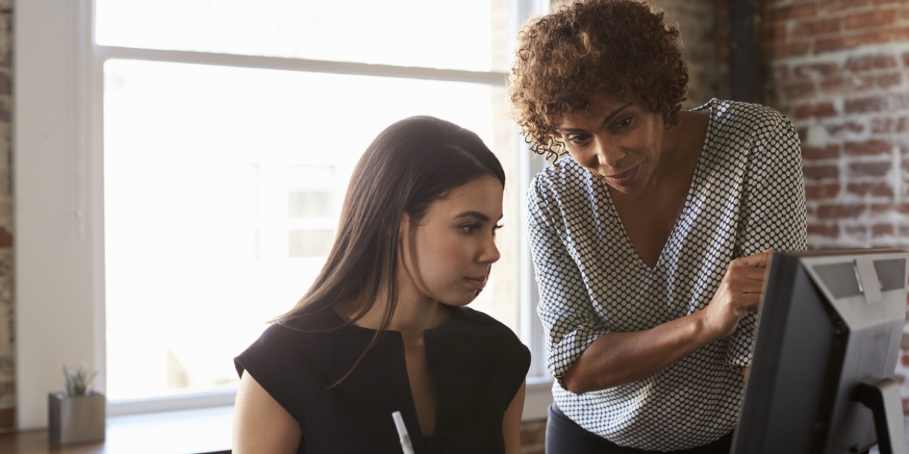 A younger woman with a black top and dark hair is sat at a computer screen. Leaning over her and also looking at the screen is an older woman wearing a spotted blouse.