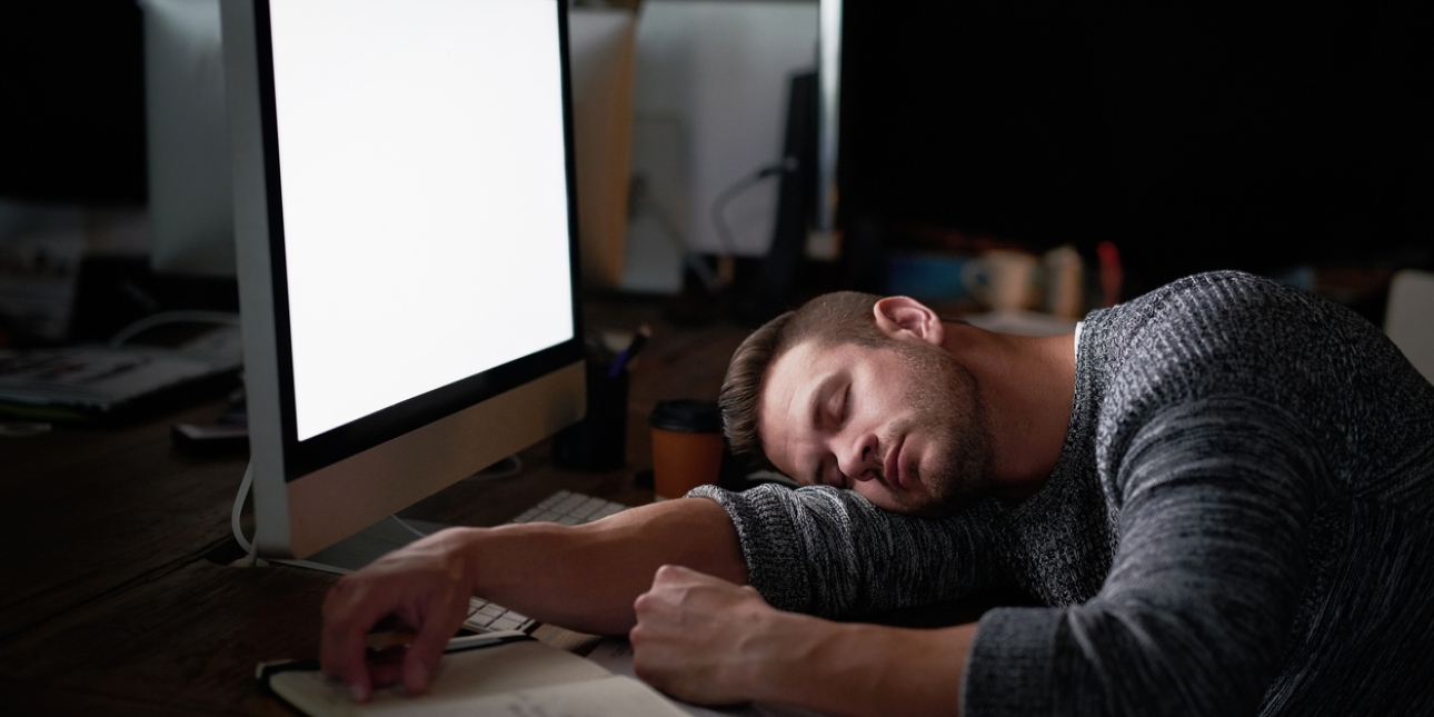 A blank computer screen lights up a dark office showing a man who has fallen asleep at his desk. Around him are a coffee cups and notebook.
