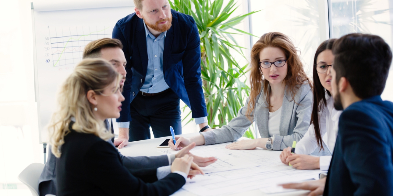 Five white business people sat at a table. A white man with ginger hair and beard is stood leaning over the table looking pensive. A woman at the table in glasses looks concerned.
