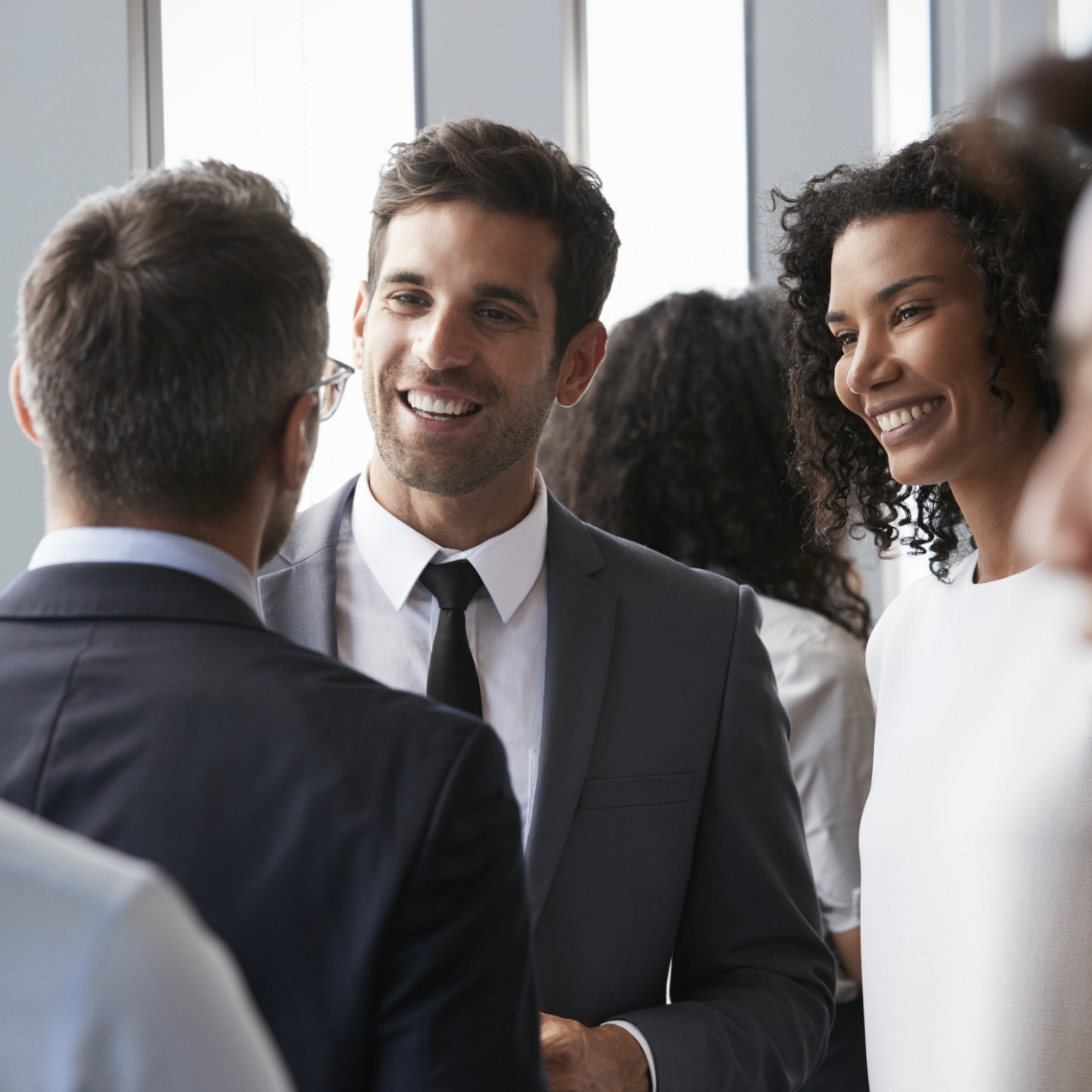 A white man with short dark hair, wearing a dark suit and tie, and a black woman with shoulder length curly hair and wearing a white blouse, smile at a business colleague at a networking event. The colleague has his back to the camera but appears in 