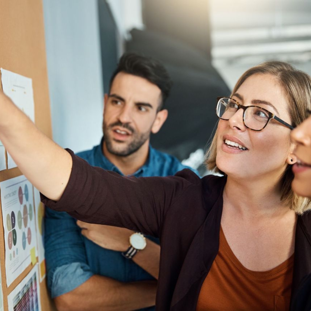 A woman in glasses reaches to a noticeboard of paper charts, while a male and female colleague, both with dark hair, look on