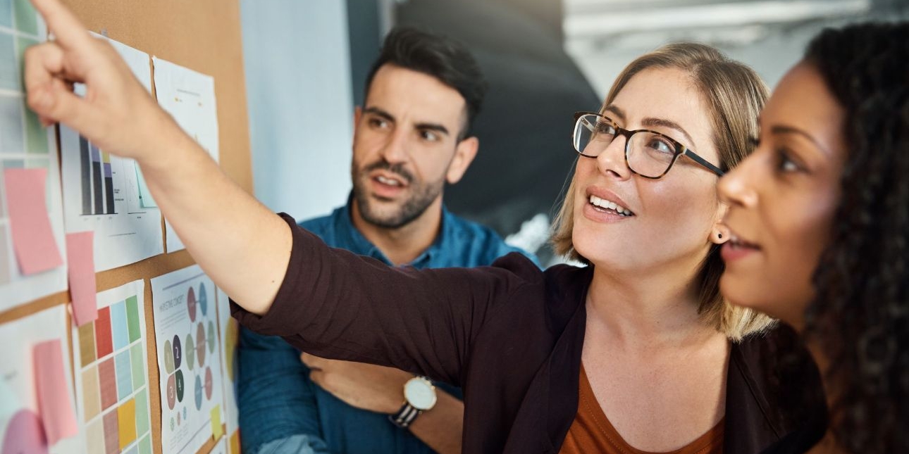 A woman in glasses reaches to a noticeboard of paper charts, while a male and female colleague, both with dark hair, look on