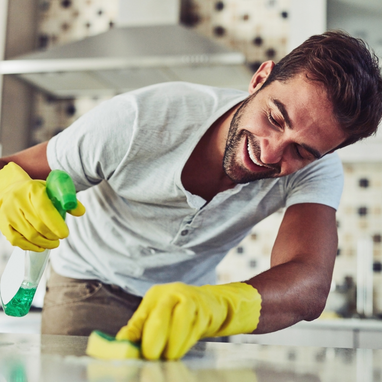 A white man with dark hair and wearing a t-shirt and yellow rubber globes holds a bottle of cleaning spray in his right hand and scrubs a kitchen surface with a scouring pad in his left hand. Behind him are kitchen units.