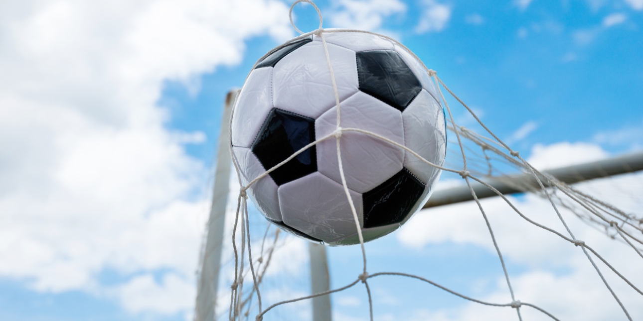 Football hits the back of the net against a bright blue but partly cloudy sky