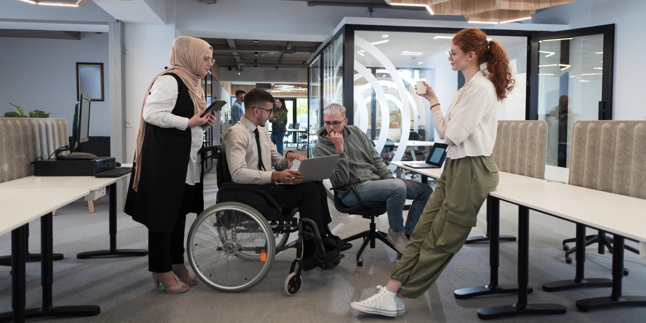 Four colleagues huddled together in a modern office. From left to right: a woman wearing black and white and a beige hijab, a man in a white shirt and dark trousers who uses a wheelchair, a man in a green hoody on a chair, a woman stood leaning again