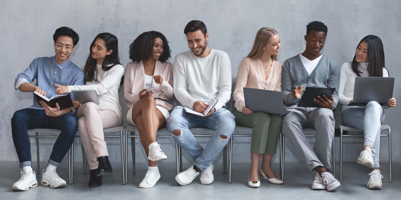 Seven casually dressed, jovial young people of different genders and ethnicities sit in a row chatting and laughing. The background is soft grey.