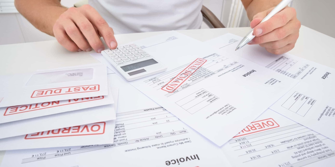 A pile of unpaid invoices and envelopes stamped with past due, final notice and overdue laid out on a desk. A pair of white hands of an unidentified person tap a calculator and hold a pen