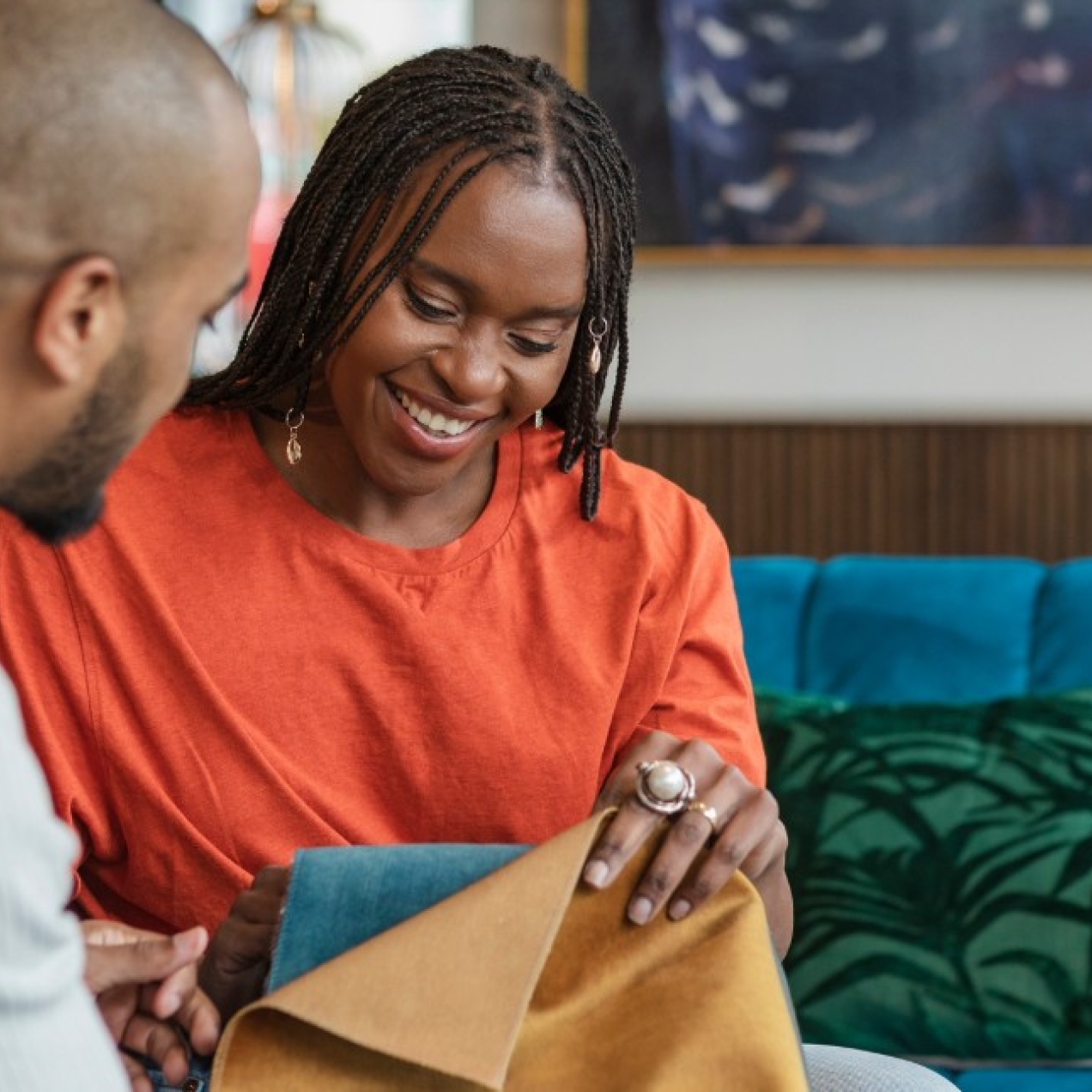 A male and female black couple sat in a furniture shop choosing sofa fabrics. They are both black; the man wears a grey top and the woman wears an orange top.