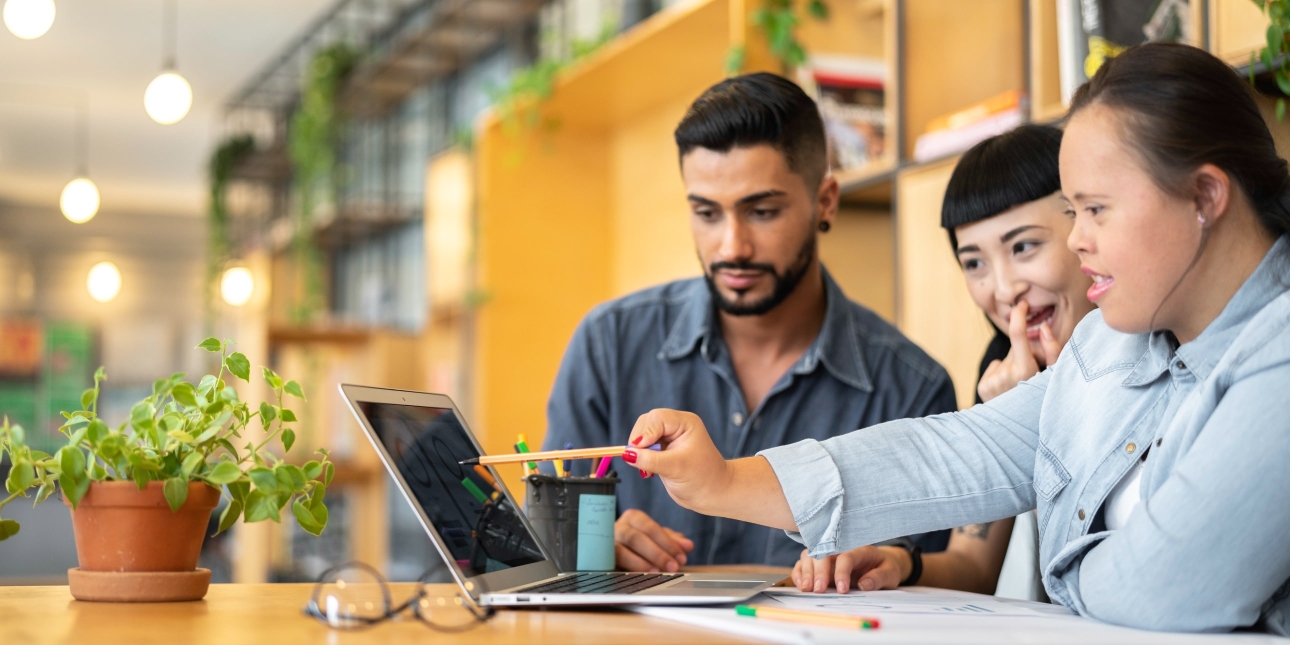 Three young, smiling colleagues gather around a laptop: an Asian man with dark hair and groomed stubble and blue shirt; an Asian woman with dark hair scratches her face; and an Asian woman with dark hair and blue shirt points a pen at a laptop screen