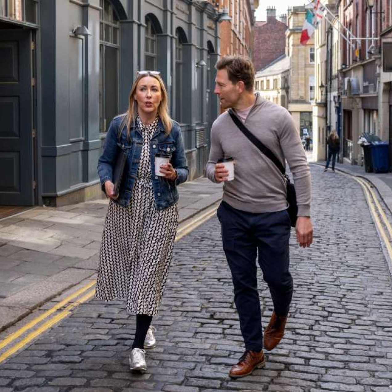 A white woman and white man in smart-casual clothing walk together along a narrow cobbled British high street while holding coffee cups.