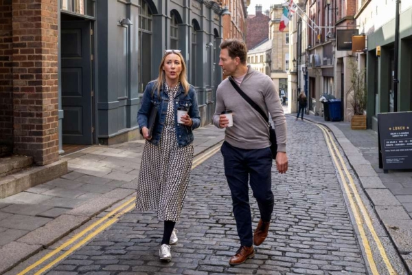 A white woman and white man in smart-casual clothing walk together along a narrow cobbled British high street while holding coffee cups.