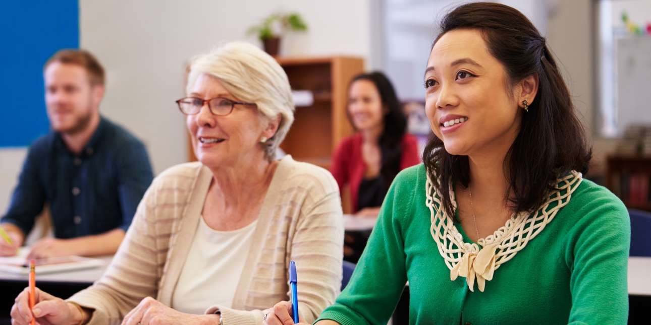 Two women sat at a desk with open notebooks in a classroom. The spectacle-wearing woman on the left is older, white, with short grey hair. The younger woman on the right is Asian, with shoulder length dark hair and wears a green top. Out of focus beh