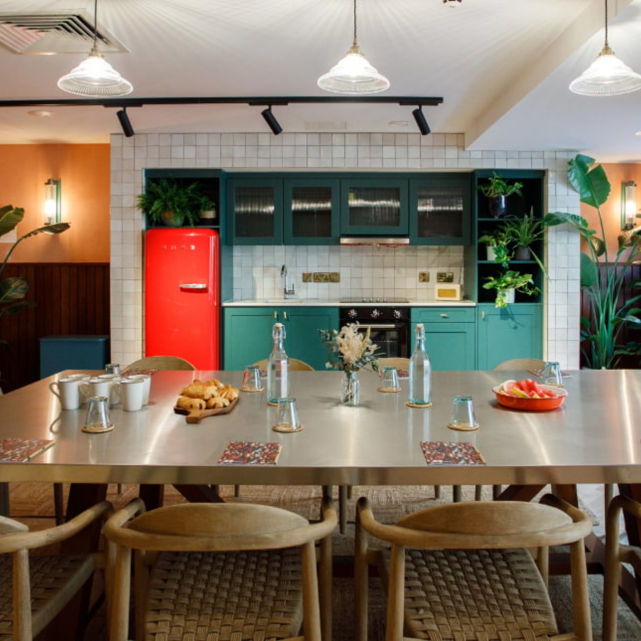 A meeting room at room2 Belfast consisting of a large light coloured table and 12 chairs, in front of a turquoise kitchen with red fridge.