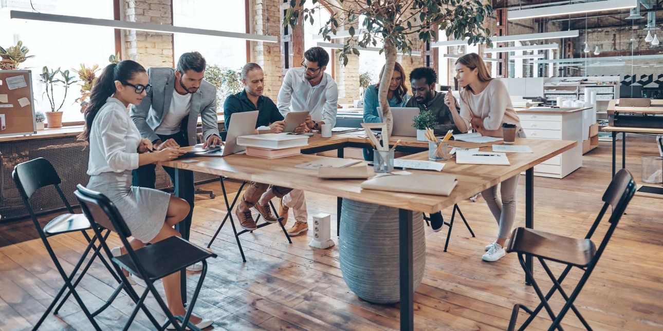 A group of six colleagues of different gender and ethnicities huddled around a table. They are concentrating on paper work and computers. There is a large plant in the middle of the desk.