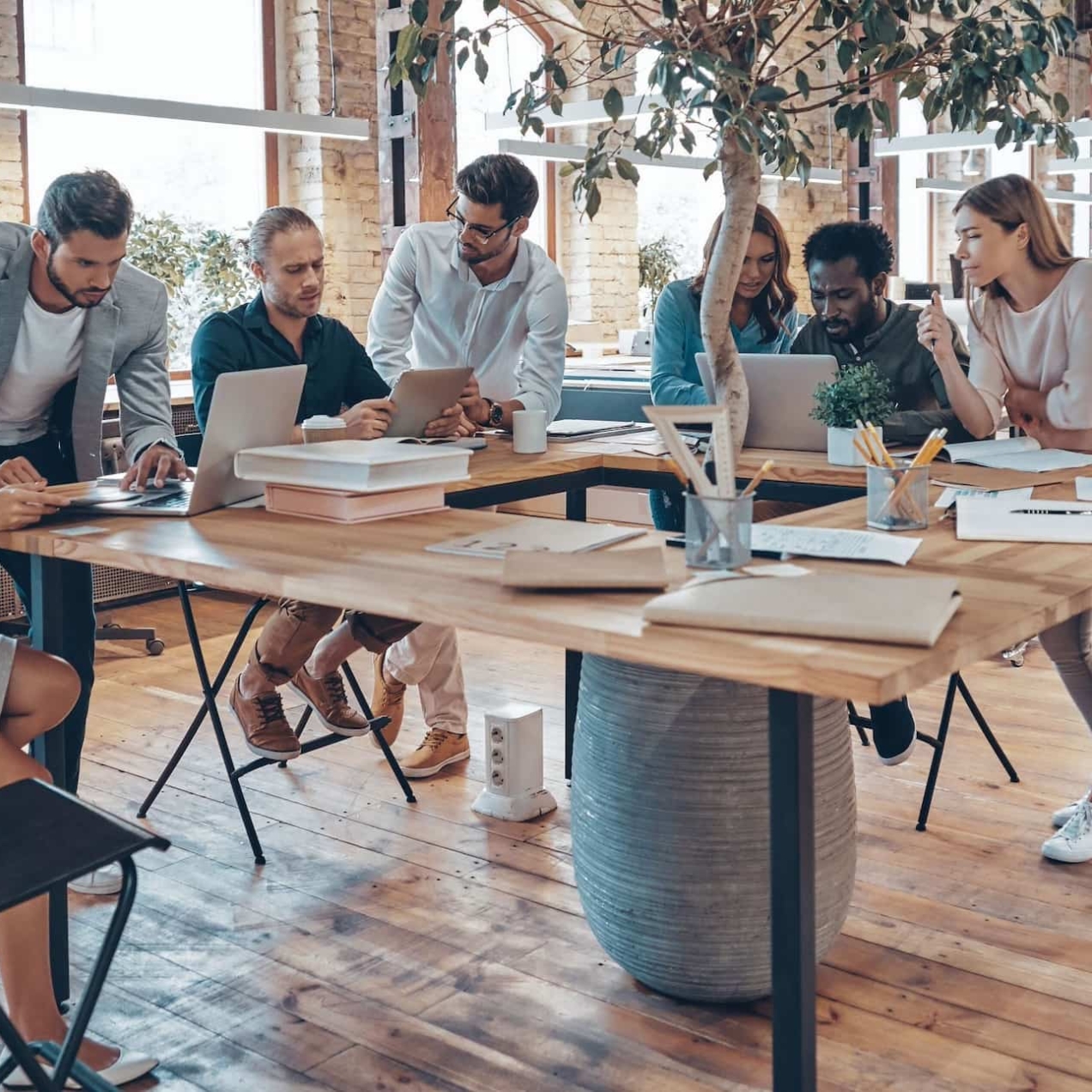A group of six colleagues of different gender and ethnicities huddled around a table. They are concentrating on paper work and computers. There is a large plant in the middle of the desk.