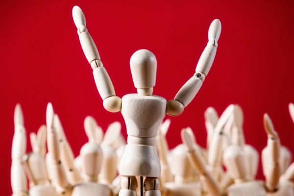 On a red background a wooden artist's mannequin stands with its arms raised addressing a crowd of other manequins.
